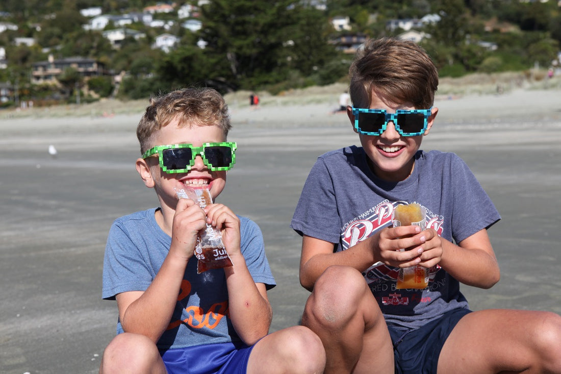 Two children eating Juicies on beach with sunglasses on in Nelson, New Zealand.